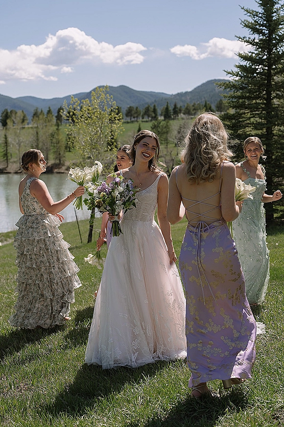 Bridesmaids with bride smiling in pastel dresses, holding calla lily bouquets in a grassy field by a lake with mountains behind