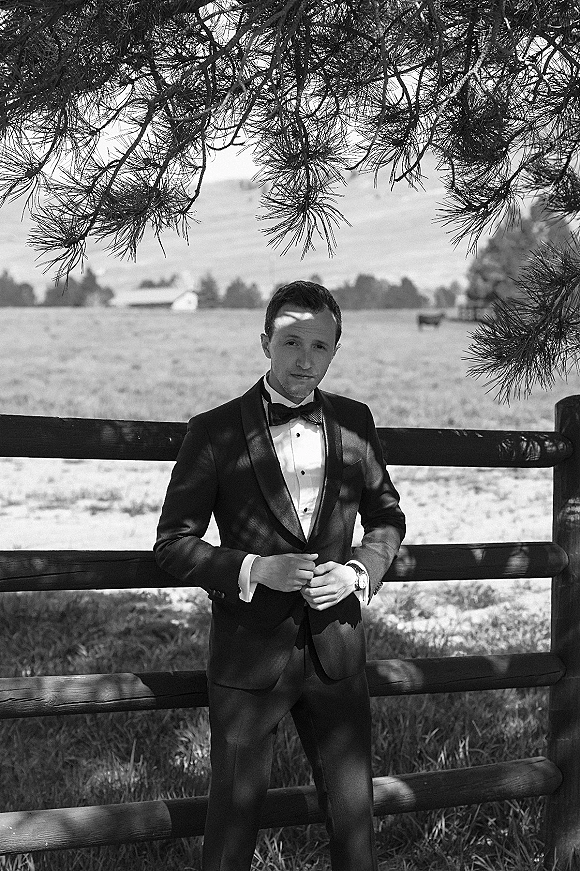 Groom portrait in a black tuxedo with bow tie, cufflinks, and wristwatch, leaning on a wood fence beside a meadow farm scene