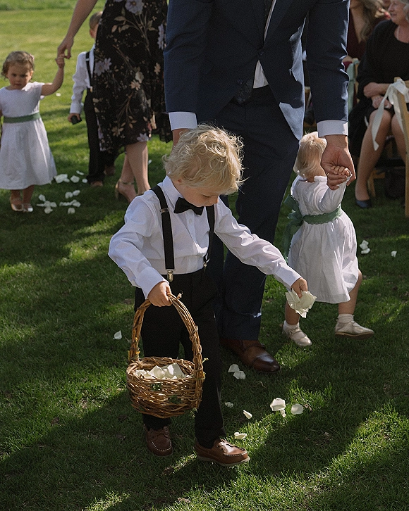 Flower boy in a flower boy outfit holds a petal basket, wearing suspenders and bow tie on an outdoor ceremony aisle with guests seated nearby