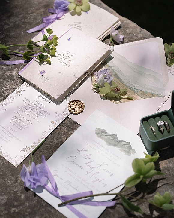 Invitation suite with vellum envelope, deckle edge paper and wax seal, styled with lavender ribbon, purple flowers, and rings on stone surface