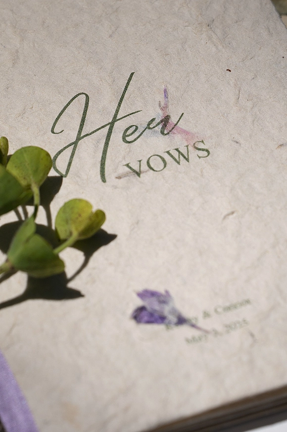 Wedding vow book with deckled edge paper and calligraphy lettering, decorated with watercolor florals and a greenery sprig on a neutral tabletop