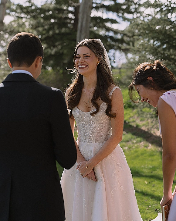 Ceremony moment as the bride laughing at altar while the groom reads vows, her veil catching sunlight on an outdoor lawn with trees