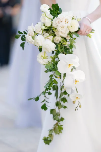 Bridal bouquet with cascading bridal bouquet orchids, white roses, and greenery held against a bridal gown, wedding ring visible, indoors