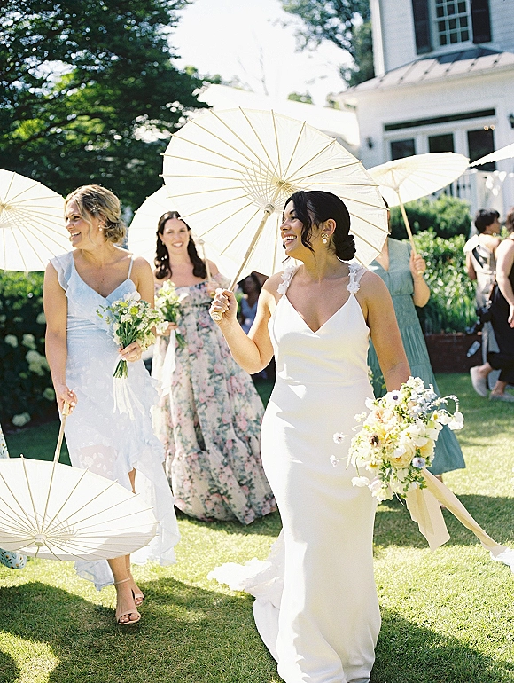 Wedding processional with bride walking under a white parasol, holding a yellow-and-white bouquet with ribbons on a garden lawn by guests