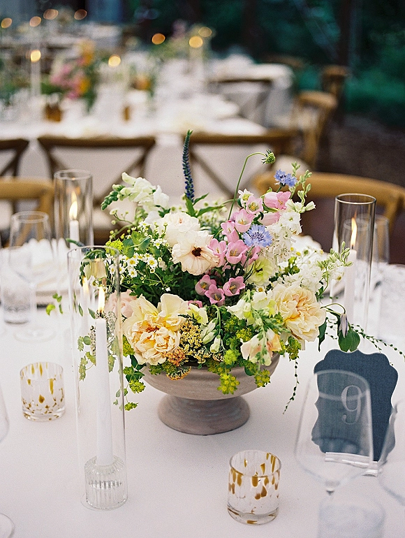 Reception tablescape with wedding table centerpiece in a compote bowl, taper candles, wine glasses, and wood chairs in an outdoor reception area