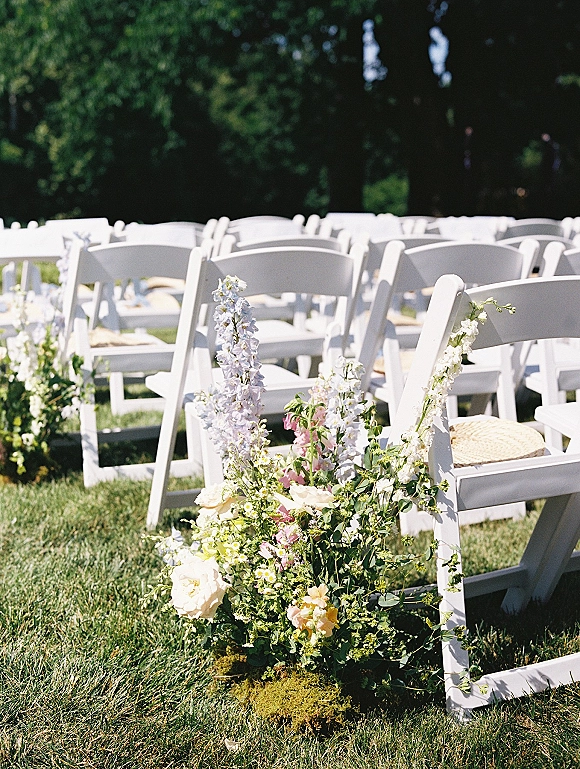 Ceremony aisle decor with outdoor wedding aisle flowers and low meadow clusters of roses, delphinium, and greenery beside white chairs on grass lawn