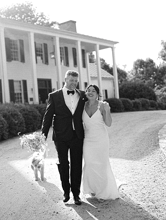 Couple portrait of bride and groom walking arm in arm with their dog, holding a bouquet on the driveway by a white columned house