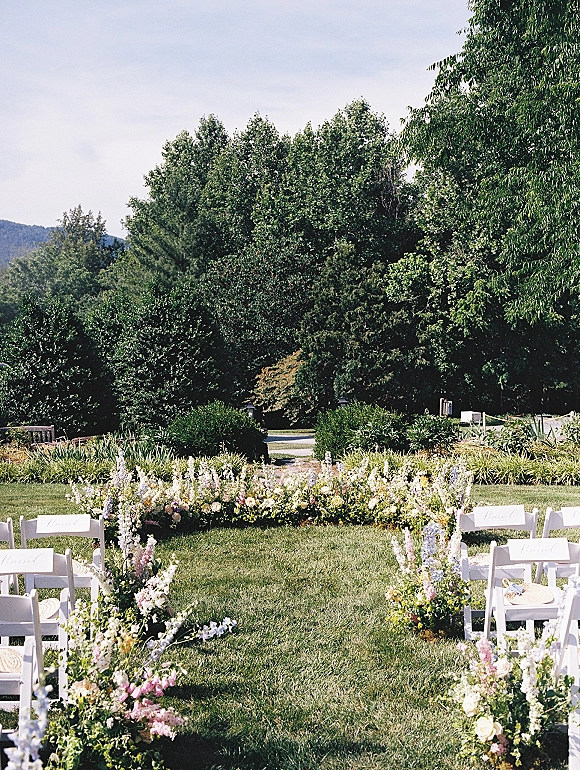 Ceremony aisle decor with white folding chairs, low pastel florals and greenery, plus woven fans and place cards on a lawn with mountains