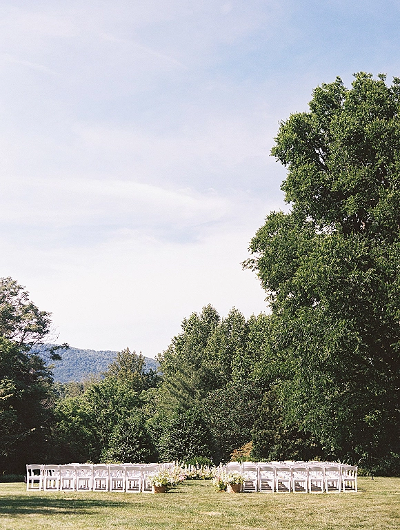 Ceremony seating with outdoor wedding chairs in neat rows, white folding chairs lining an aisle with basket florals on a green lawn and hills beyond