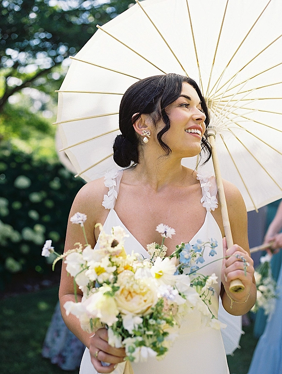Bridal portrait of a bride with parasol holding a white and blue wildflower bouquet, looking away in a garden with blurred bridesmaids behind