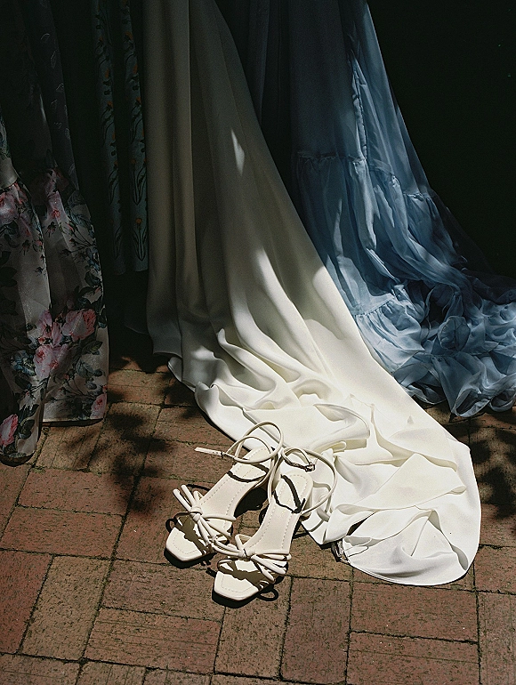 Bridal shoes, white strappy bridal heels with bow detail on a brick patio beside a wedding dress train with hanging dresses above