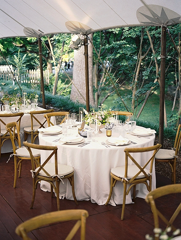 Reception tablescape with outdoor tent reception style—round table in white linen, cross back chairs, floral centerpiece and candlelight under a clear top tent
