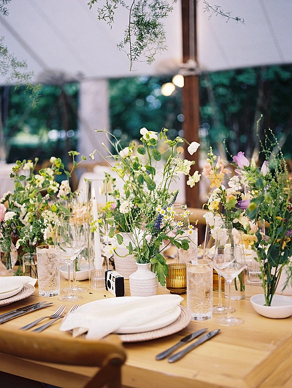 Reception tablescape with wildflower wedding centerpieces in bud vases, taper candles and amber votives on a long table under string-lit tent canopy