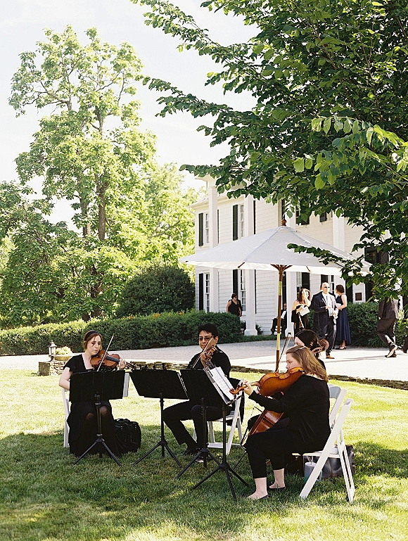 Wedding musicians performing as a wedding string quartet with music stands and sheet music on a green lawn near a white house exterior
