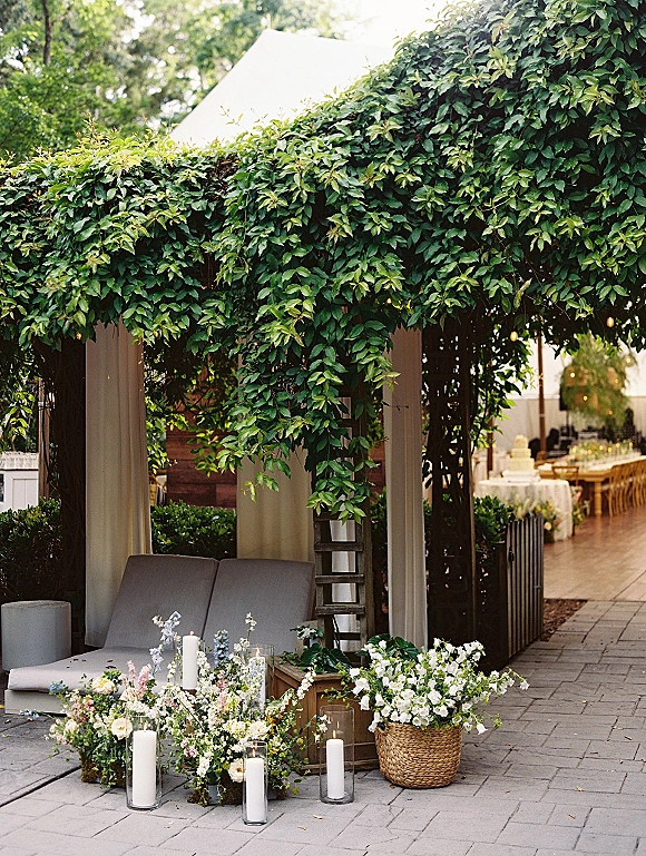 Outdoor lounge decor with wedding lounge seating under a vine-covered pergola, cabana curtains, sofa, and white pillar candles on a patio
