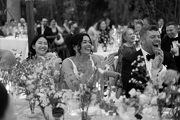 Wedding reception guests in tuxedos and bridesmaid dresses laughing at banquet tables with bud vases and floral centerpieces under trees