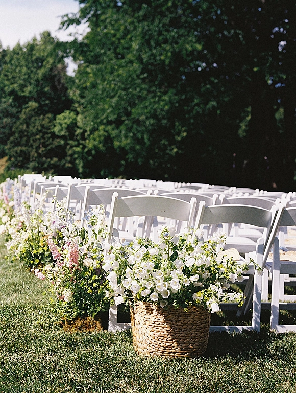 Ceremony seating with outdoor ceremony chairs in neat rows, aisle lined with white and pink florals and a wicker basket planter on a lawn under trees
