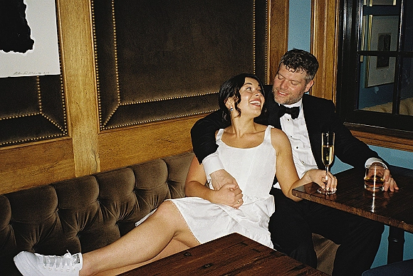 Couple portrait of bride in a short wedding dress and groom in tuxedo cuddling in a tufted booth, toasting champagne and whiskey