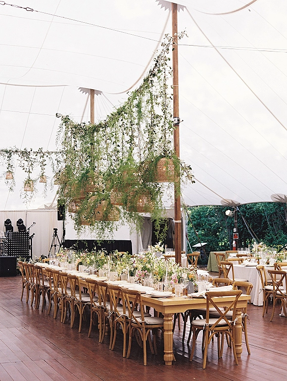 Reception tablescape with a long banquet table setup, white linens, pastel bud vases and greenery garland under a white tent with string lights
