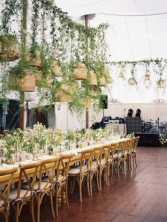 Reception tablescape with a long banquet table wedding setup, floral bud vases and taper candles under hanging wicker lanterns in a white tent