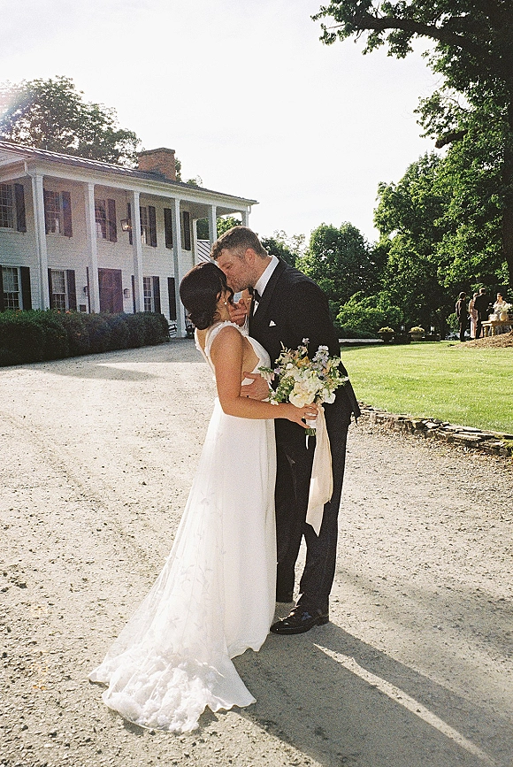 Wedding kiss portrait of bride and groom kissing as she holds a bouquet with trailing ribbon, outside a historic house with columns and guests behind