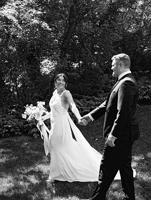 Couple portrait of bride and groom holding hands, bride looking back with bouquet ribbon, in dappled sunlight among garden trees.