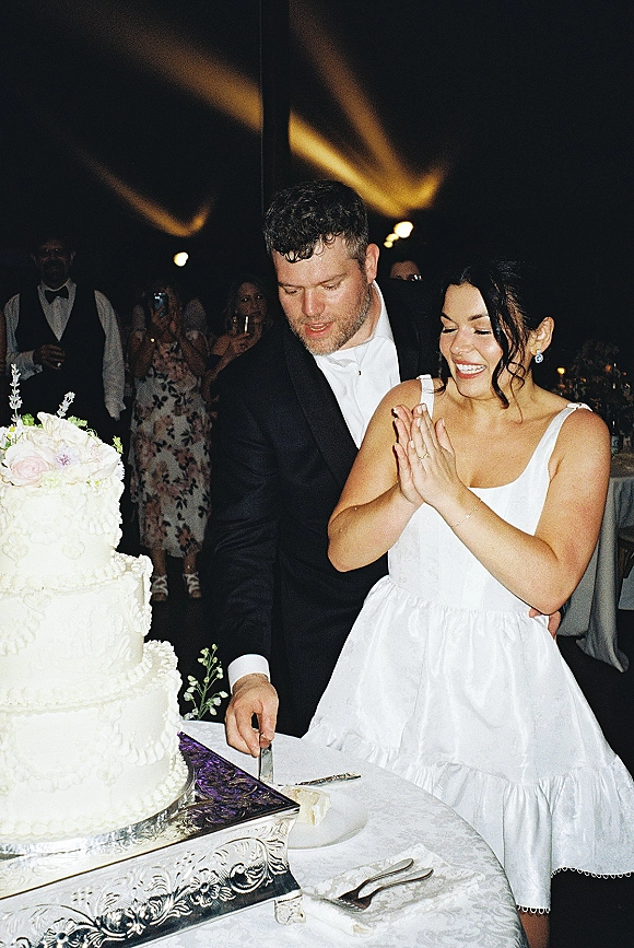 Wedding cake cutting as bride in a white dress and groom in black suit slice a three-tier cake under string lights in a tented reception