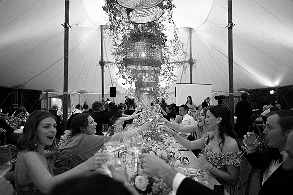 Wedding reception toast as guests raise wine glasses along a long banquet table with floral runner, taper candles, under a sailcloth tent