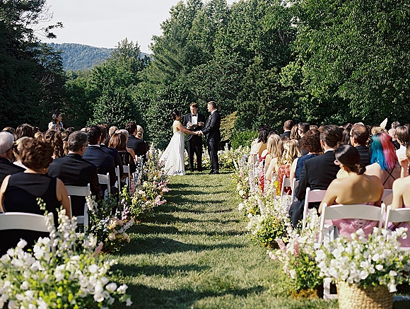 Outdoor wedding ceremony with bride and groom at the altar, garden wedding ceremony seating with white chairs and basket aisle flowers on a green lawn