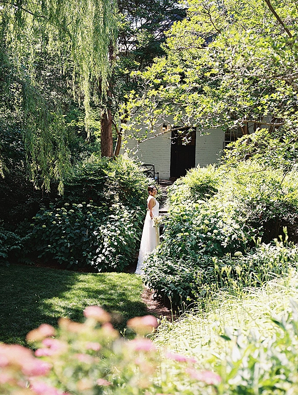 Bridal portrait of a bride holding bouquet, looking back on a garden path in a classic white gown with greenery and a white brick building behind
