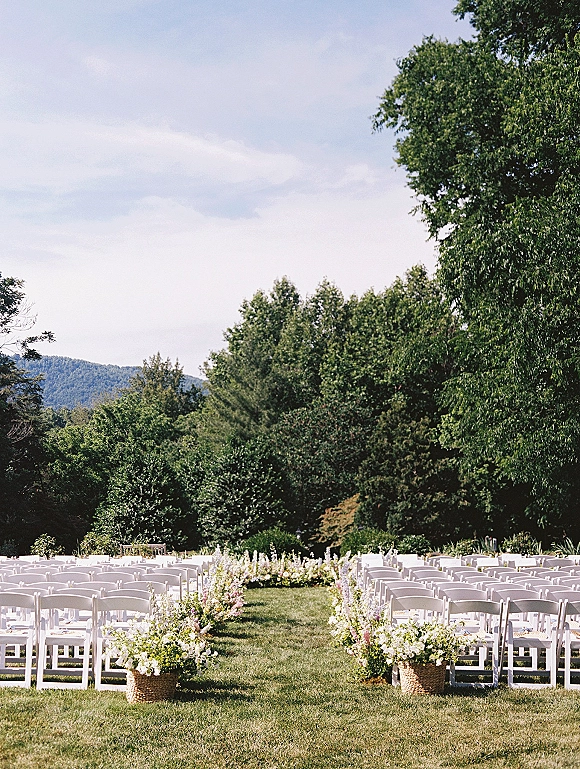 Outdoor ceremony setup with white folding chairs and wicker basket aisle flowers on a lawn, framed by trees and mountain views