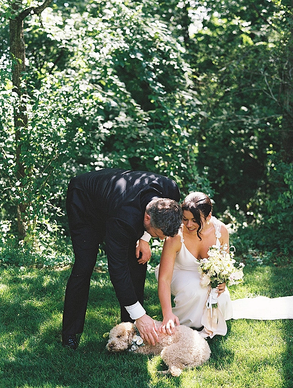 Wedding couple with dog kneeling on green grass, bride in veil holding bouquet as they pet their pup with a floral collar in dappled sunlight