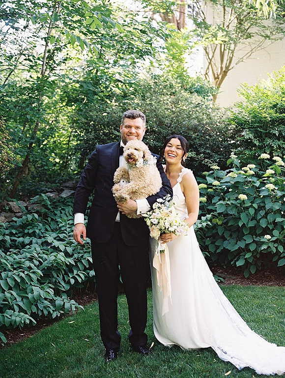 Couple portrait of bride in a simple wedding dress and groom in a black tuxedo holding their dog with floral collar on a garden lawn