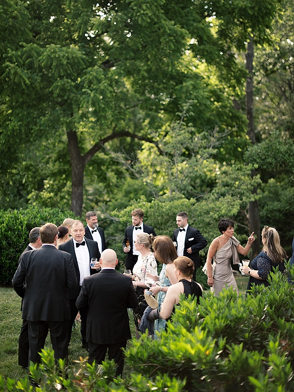 Wedding cocktail hour guests mingle with outdoor cocktail hour drinks in tuxedos and formal dresses on a garden lawn with trees and greenery