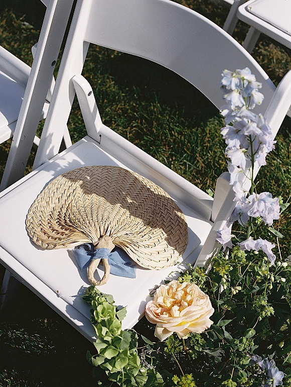 Ceremony chair decor with a woven hand fan tied by a blue ribbon to a white folding chair, accented with peach rose blooms on a lawn