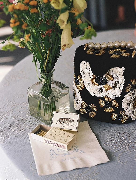 Wedding details flat lay of bridal accessories with black beaded clutch, pearl handle, matchbox, napkin, and flowers on lace tablecloth