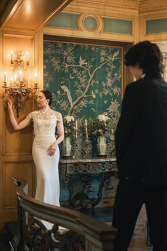 Bridal portrait of a bride in a high neck wedding dress with lace bodice, posed by chinoiserie wallpaper near taper candles and florals