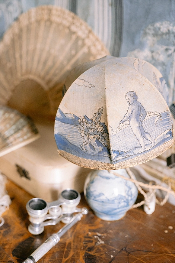 Vintage wedding details in a wedding flat lay with a hand-painted blue-and-white lamp, opera glasses, fan, and books on wood tabletop