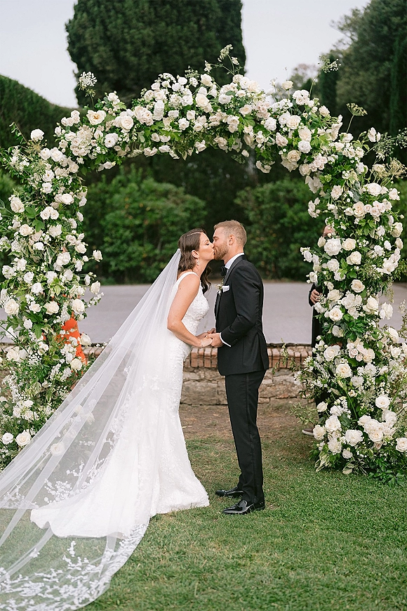 Wedding kiss portrait of bride and groom kissing under a white rose floral arch, her long veil and lace dress flowing in a garden setting