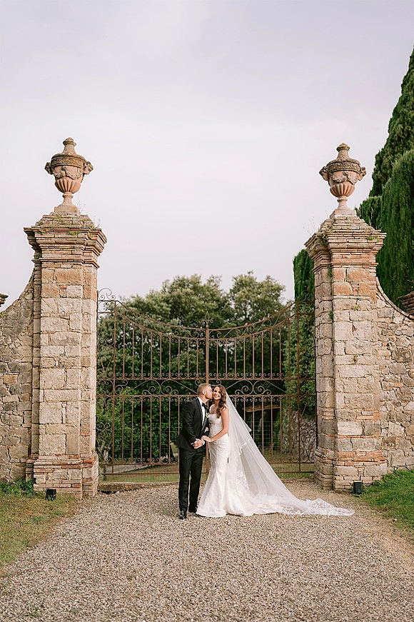 Couple portrait of bride and groom kiss, holding hands at a wrought iron gate with stone pillars; her long veil drapes behind them