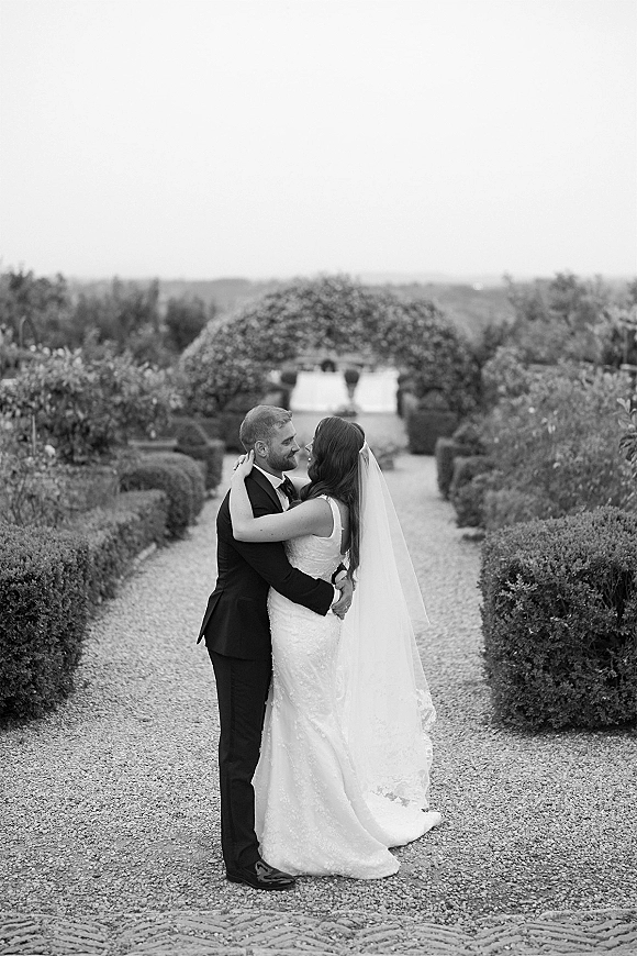 Couple portrait in a black and white wedding portrait style, bride in lace dress and long veil embracing groom on a garden path with hedges