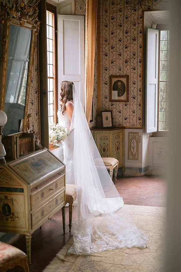 Bridal portrait of a bride at window in a lace wedding dress with long veil train, holding a white rose bouquet in a vintage room