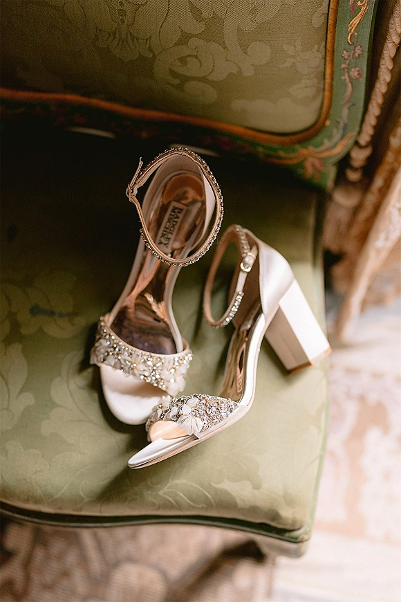 Wedding shoes with rhinestone embellishments and ankle straps resting on a vintage upholstered chair with patterned green fabric in soft light