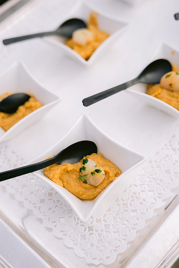 Wedding appetizer cups with herb garnish in white square bowls and black plastic spoons, arranged on a white serving tray with lace doily