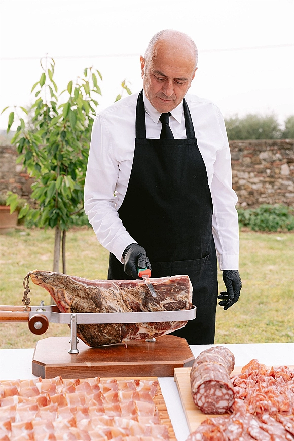 Wedding catering chef in black apron and gloves carving a ham leg at a charcuterie station wedding on an outdoor lawn by a stone wall