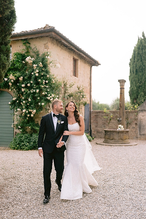 Couple portrait of bride and groom walking hand in hand, her strapless dress train flowing in a stone villa courtyard with roses