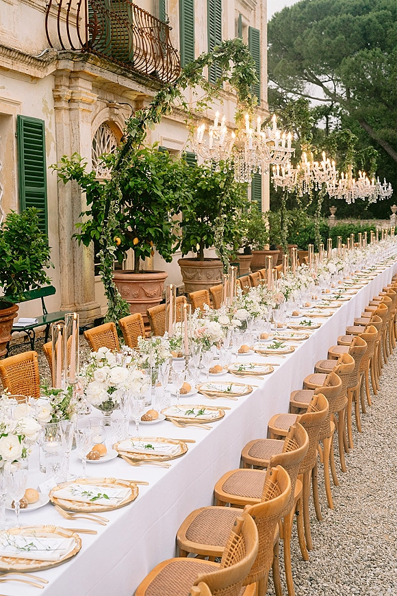 Reception tablescape with a long wedding banquet table, white linen, greenery garland, candles, and chandeliers in a stone villa courtyard