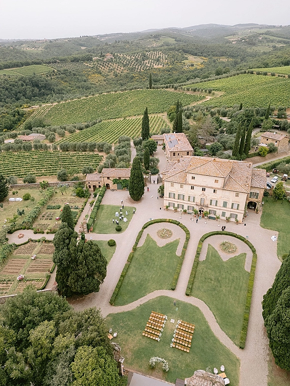 Wedding venue aerial view of a stone villa and lawn ceremony chairs with aisle florals, set amid vineyard hills and olive groves