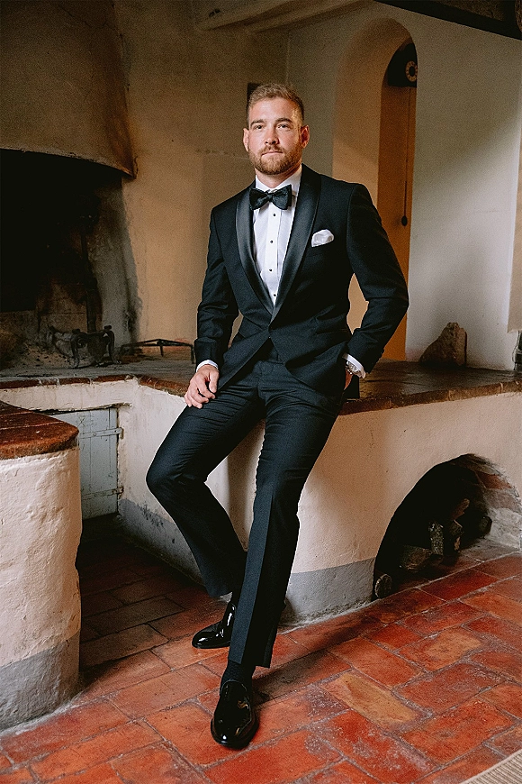 Groom portrait in a black tuxedo groom look with bow tie and pocket square, posed by an arched fireplace on terracotta tile floor