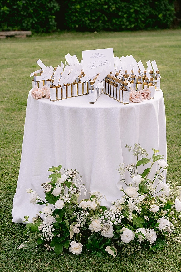 Wedding favors table with mini bottle wedding favors and escort cards on a white linen cocktail table, with rose and greenery accents on a lawn